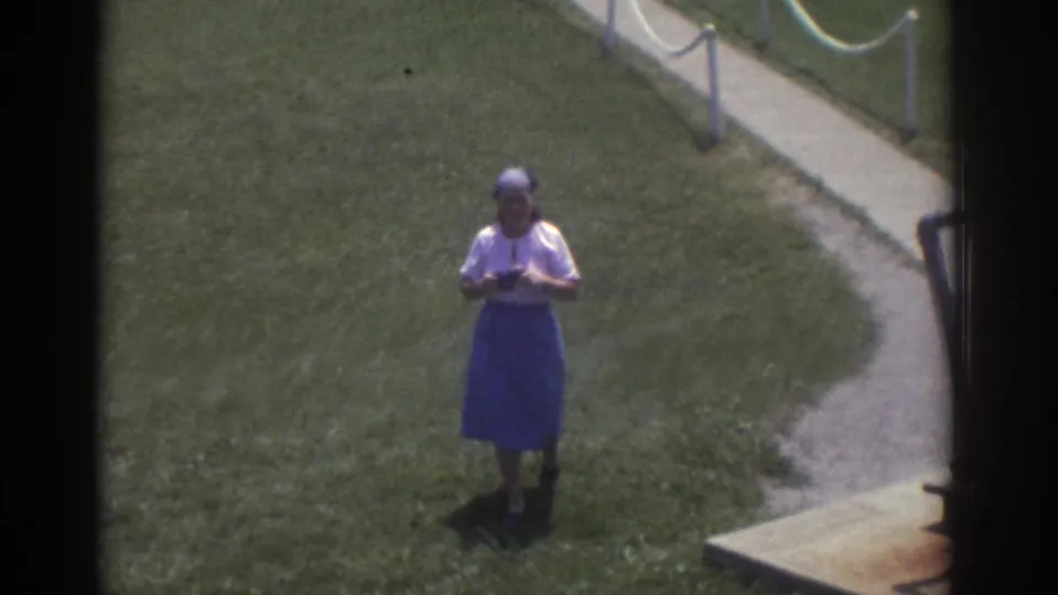 The woman lowers or adjusts her camera while standing on grass beside a curved walkway in the 1974 Cleveland visitor scene.