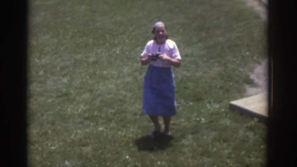 Closer 8mm view of the woman in a headscarf, white blouse, and blue skirt holding a camera on the lawn in Cleveland.