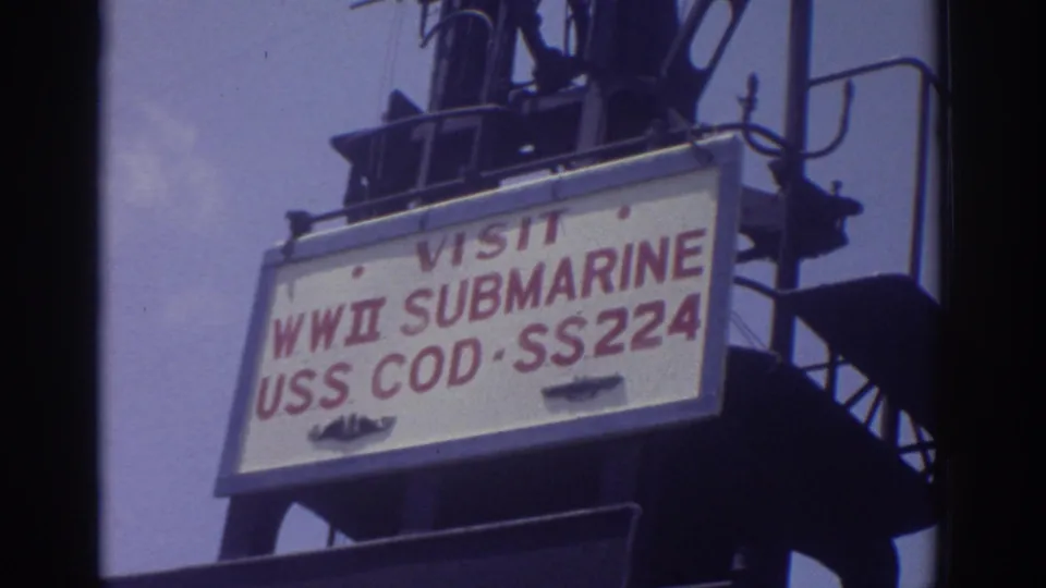 1974 Cleveland home-movie frame showing the USS Cod SS224 visitor sign mounted high on the submarine tower against sky.