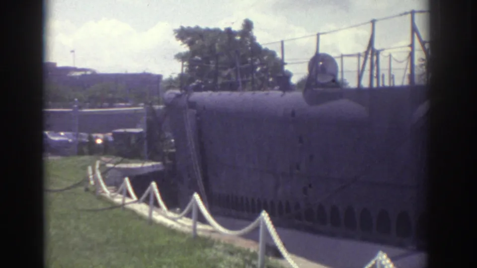 Visitor-shot 1974 Cleveland view of the submarine display area, with rope posts, chain-link fencing, grass, trees, and parked cars nearby.