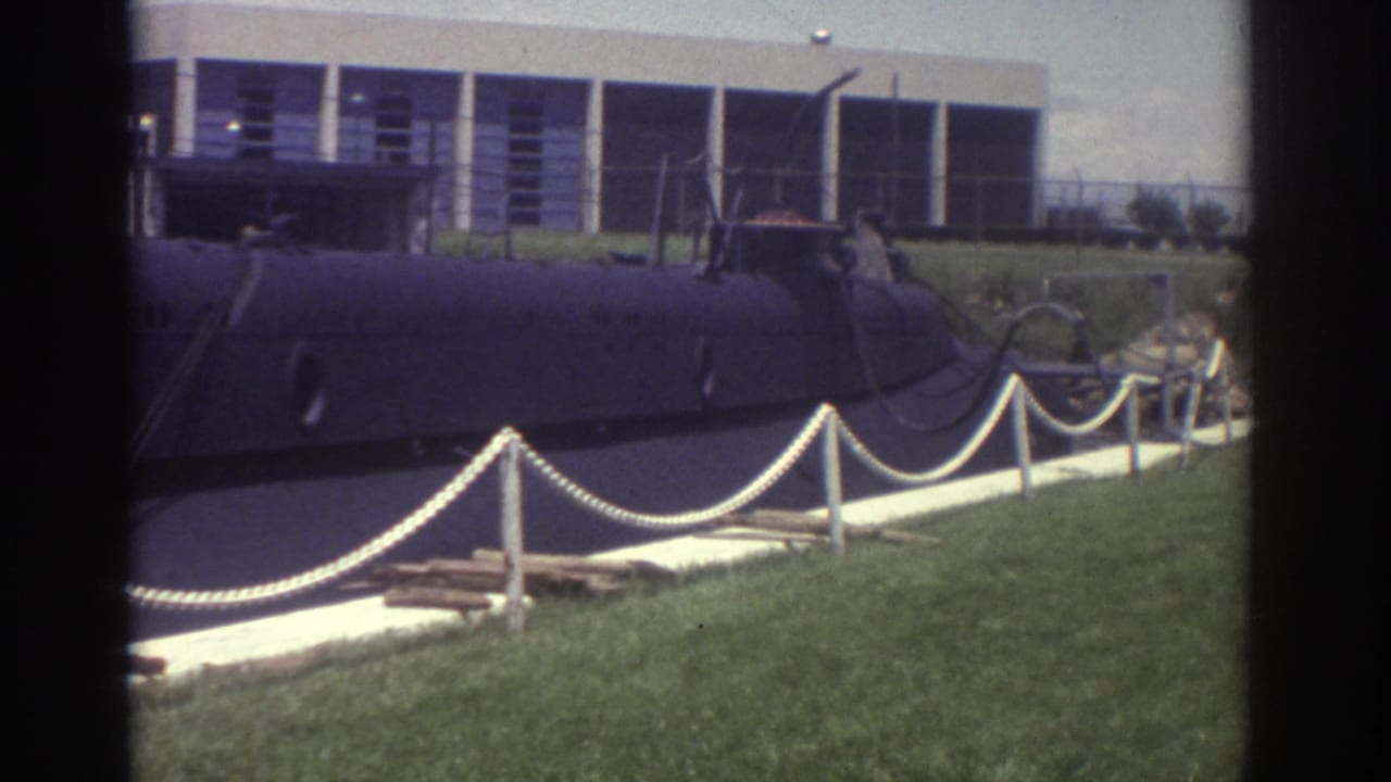 1974 Cleveland home-movie frame showing a dark submarine-like vessel on display beside lawn, rope barriers, and a modern building.