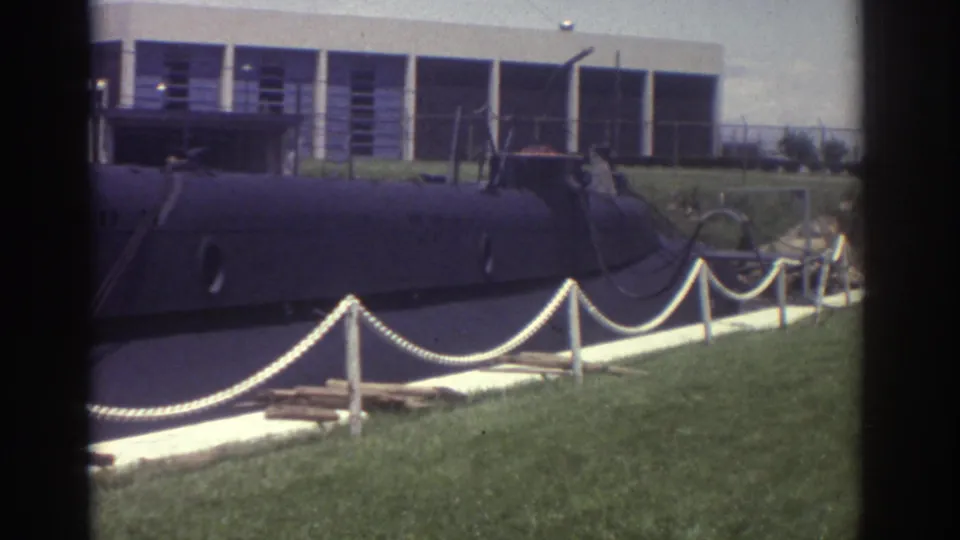 1974 Cleveland home-movie frame showing a dark submarine-like vessel on display beside lawn, rope barriers, and a modern building.