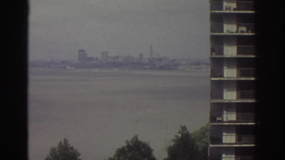 1974 Cleveland home-movie frame looking from a high-rise hotel window across a large body of water toward a hazy distant skyline, with a neighboring building edge in view.