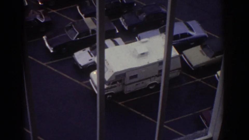 Overhead 1974 Cleveland hotel-window view of a marked parking lot with several 1970s cars and a prominent white camper van below.