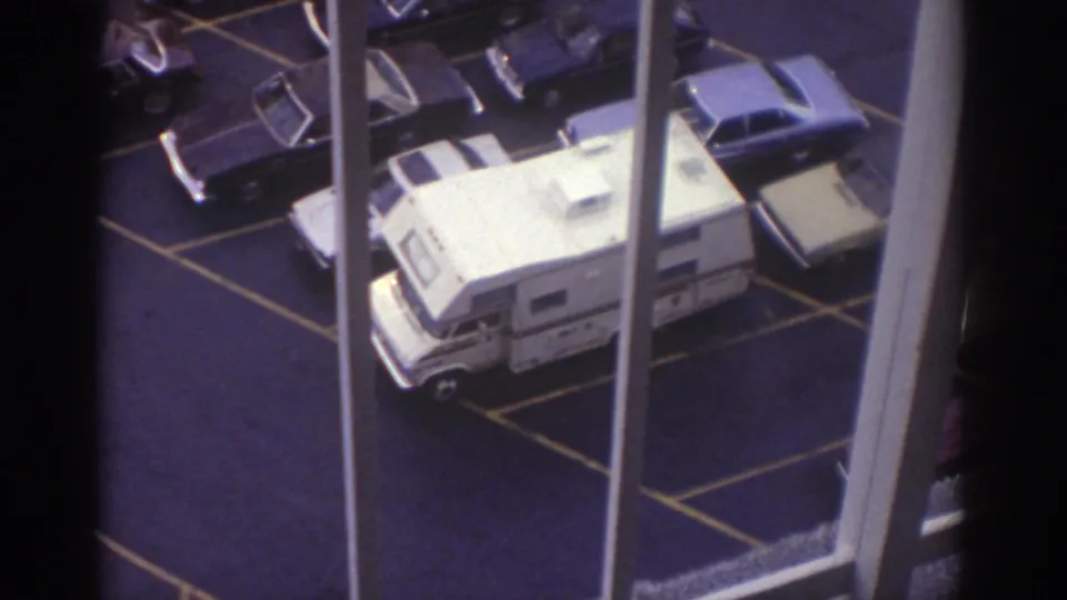 High-angle 8mm frame from a Cleveland hotel window showing a white camper van surrounded by parked cars in a striped parking lot.