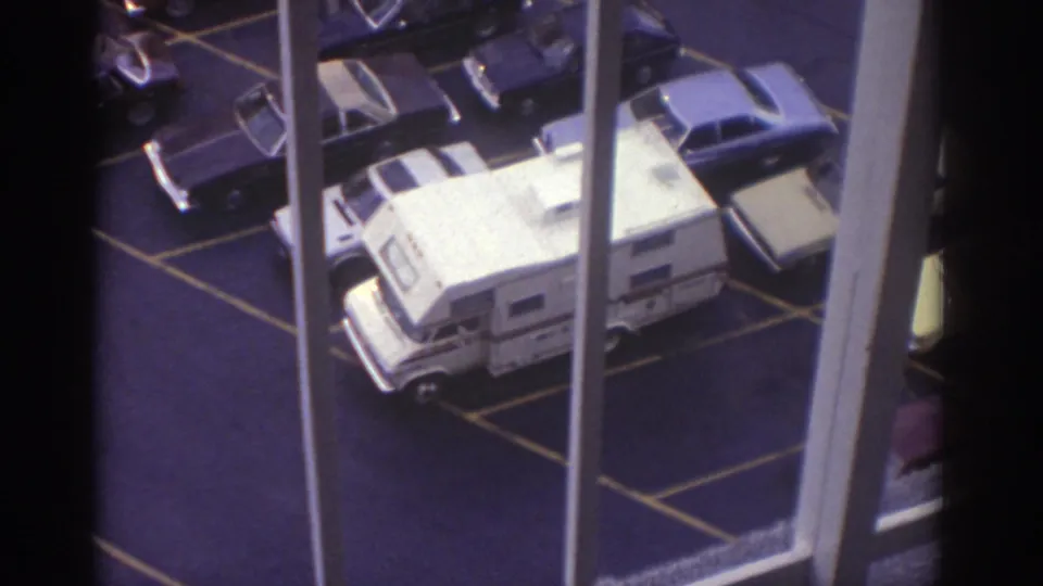 1974 Cleveland home-movie view looking down through hotel-window framing at a white camper van, surrounding parked cars, and painted parking spaces.