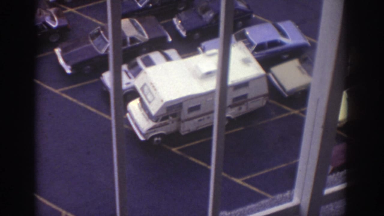 1974 Cleveland home-movie frame looking down at a parking lot with a white motorhome, classic cars, painted lines, and vertical window bars.