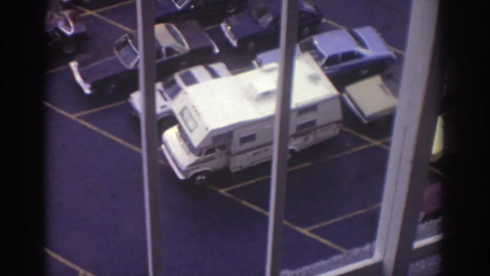 1974 Cleveland home-movie frame looking down at a parking lot with a white motorhome, classic cars, painted lines, and vertical window bars.
