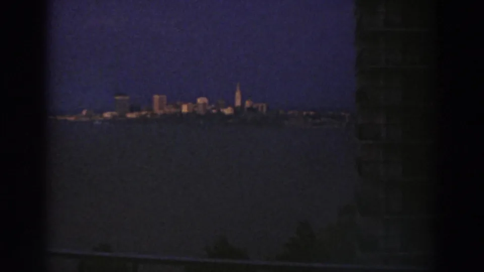 Distant Cleveland skyline across a dark waterway at dusk, with lit buildings, a tall spire, deep blue sky, and a dark foreground edge.