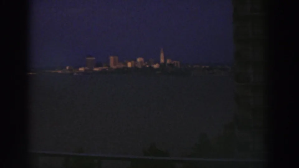 Wide 1974 Cleveland skyline view across a dark waterway, framed by shadowed edges and a dim foreground in blue dusk light.