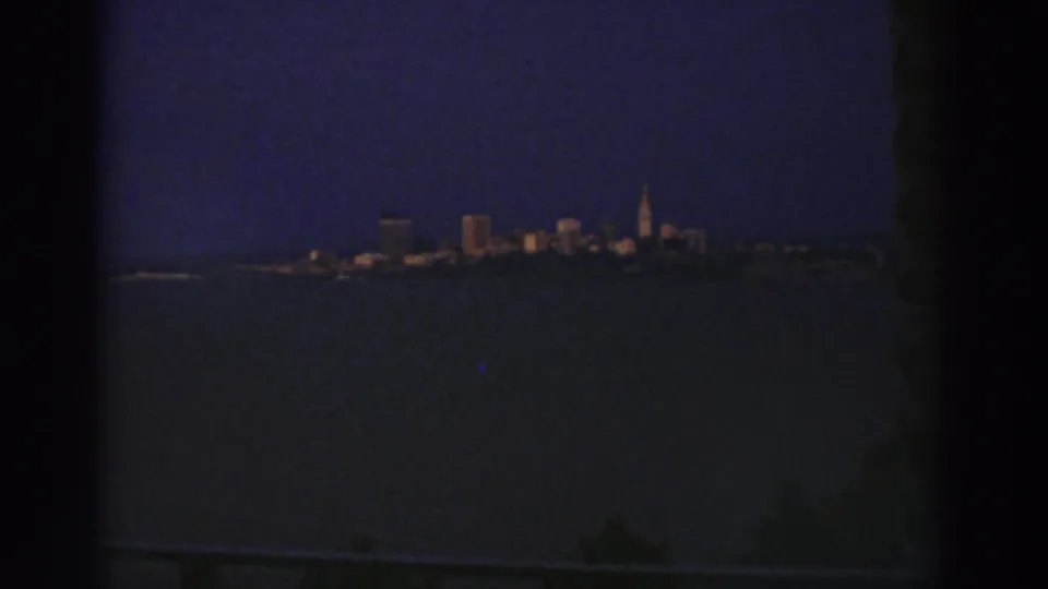 Cleveland skyline across a dark waterway at dusk, showing scattered lit buildings, a prominent spire, and a dark structure at the frame edge.
