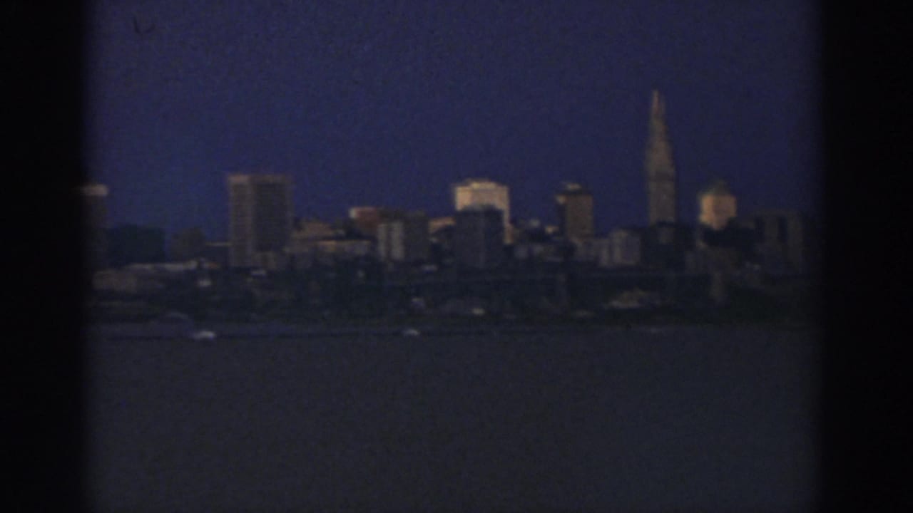 1974 Cleveland skyline seen across dark water, with a tall spire and soft 8mm grain at dusk or night.