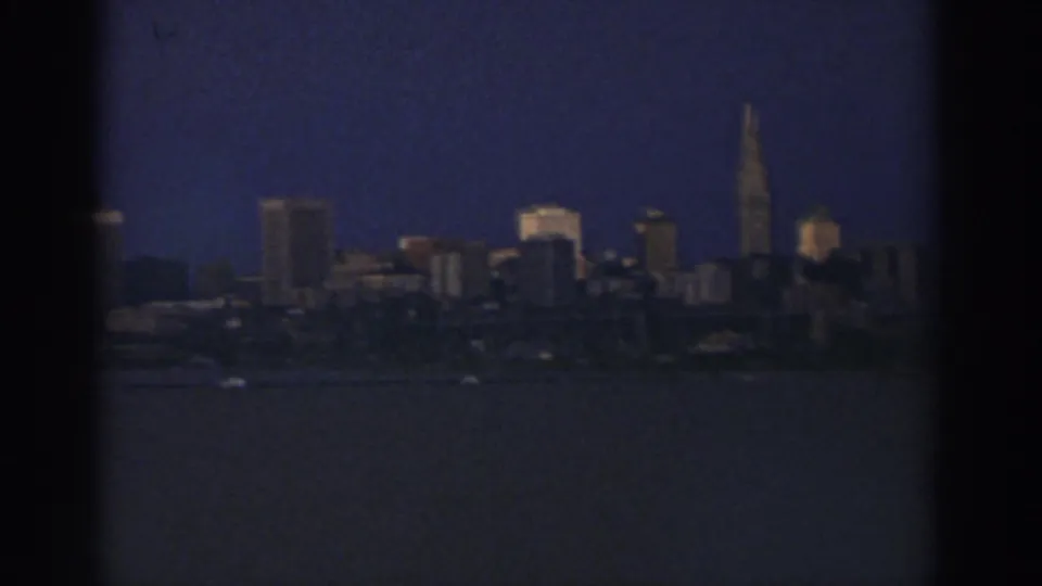 1974 Cleveland skyline seen across dark water, with a tall spire and soft 8mm grain at dusk or night.