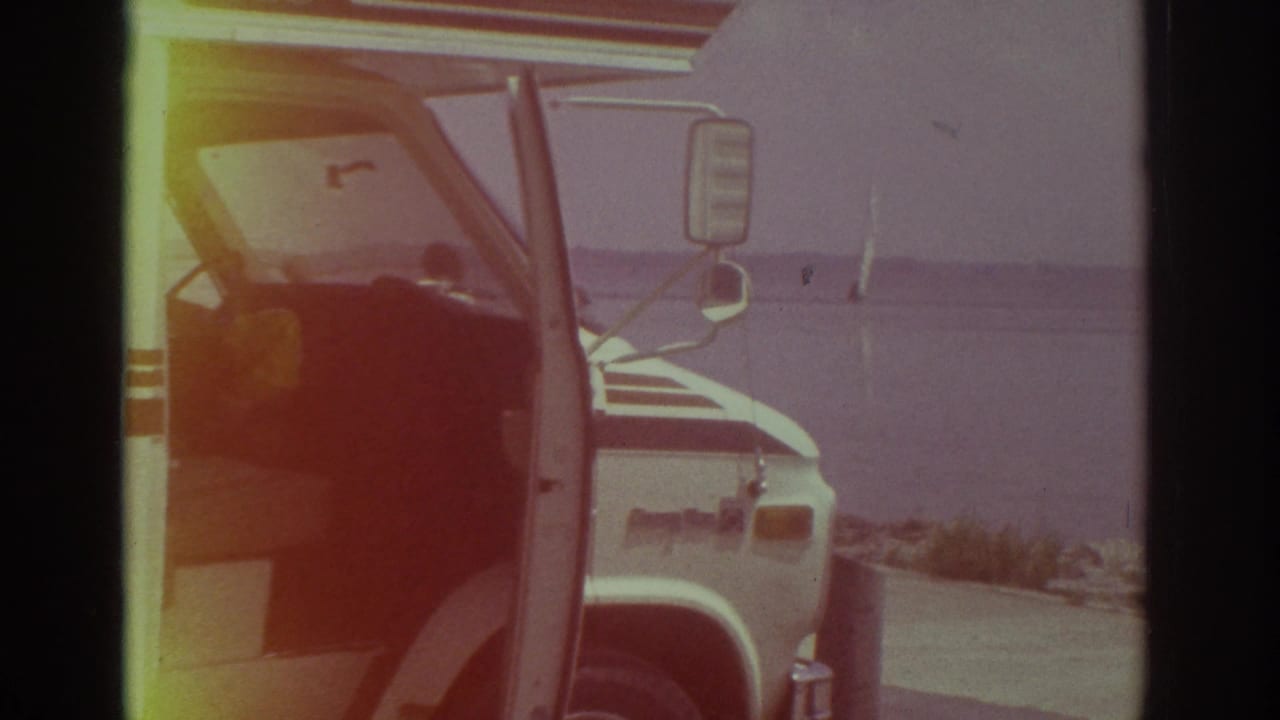 1974 Cleveland home-movie frame showing a white camper van parked near a shoreline, its door open, with water and a sailboat beyond.