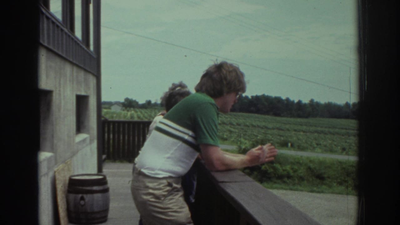 Adults leaning on a wooden balcony railing beside a building, looking over a green field.