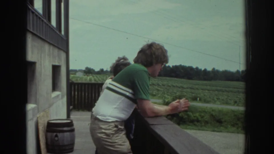 1974 Cleveland home-movie frame of adults on a balcony beside a building, looking across a green field from a wooden railing.