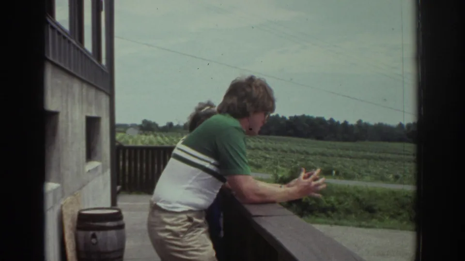 1974 Cleveland home-movie frame showing a man in a green and white striped polo leaning at a balcony rail above open fields.