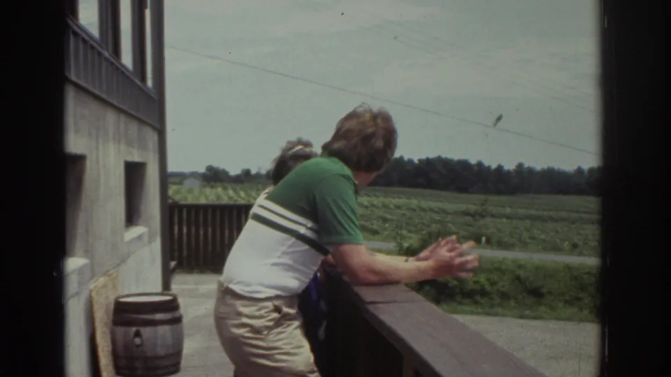 1974 Cleveland home-movie frame of a man leaning on a wooden balcony railing, with another adult partly visible and fields beyond.