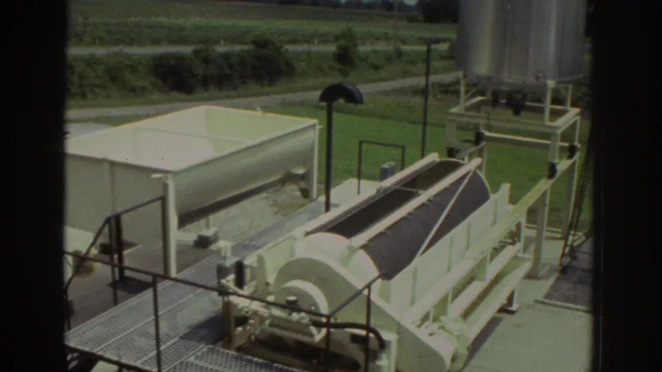 1974 8mm view of industrial processing equipment with a white tank, covered drum assembly, railings, and rural grassland.
