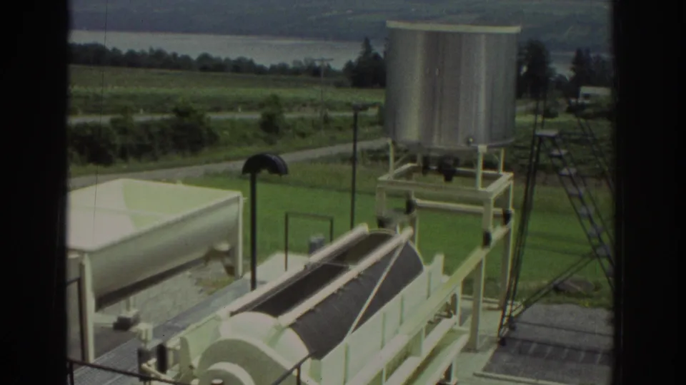 1974 Cleveland-area home-movie frame showing plant equipment, a tall stainless tank, ladder, road, and surrounding fields.