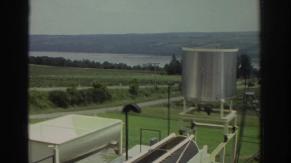 1974 8mm frame from the Cleveland-area clip showing industrial tanks against fields, distant water, and low hills.