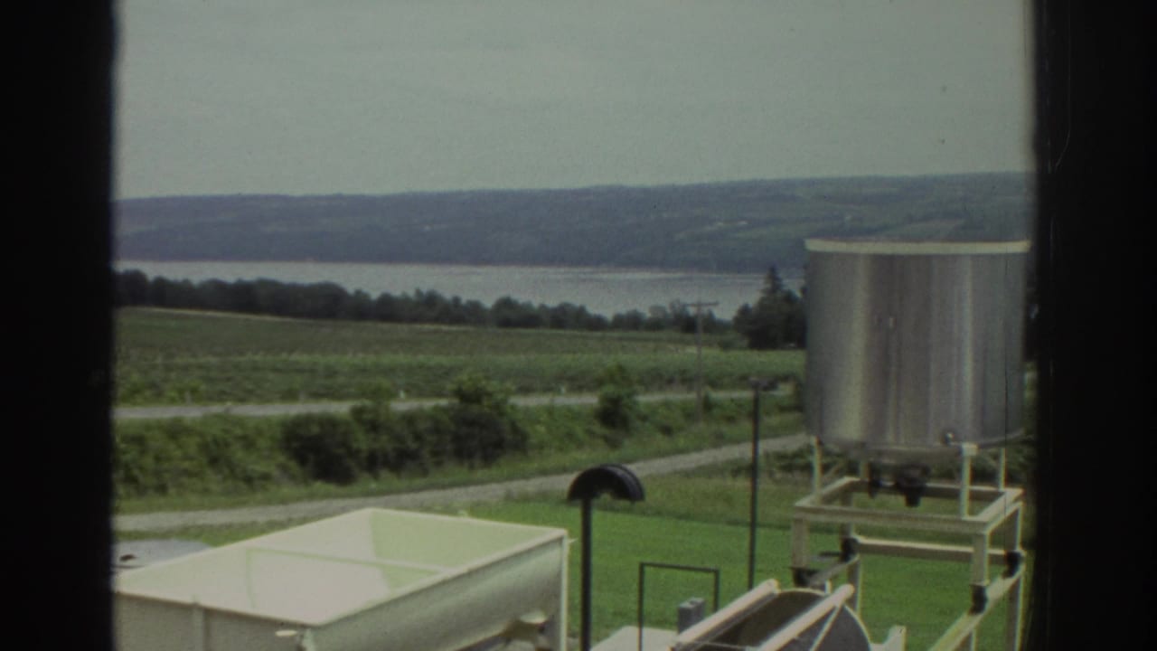 1974 Ohio home-movie view through a window showing a silver tank, pale rectangular vessel, fields, road, and distant water.