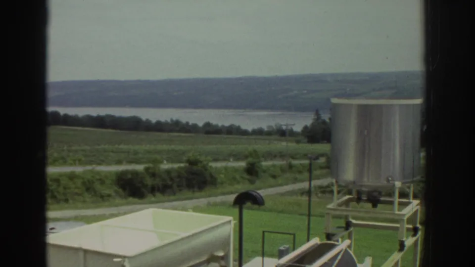 1974 Ohio home-movie view through a window showing a silver tank, pale rectangular vessel, fields, road, and distant water.