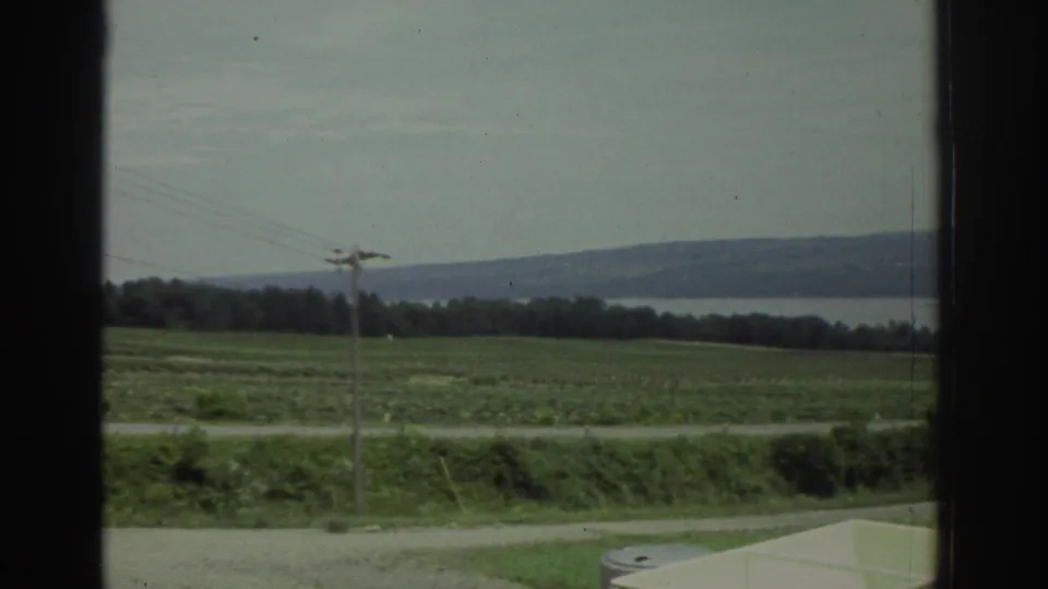 Cautious 1974 Ohio home-movie frame of a field, road, utility pole, distant water, hills, and a pale tank corner.