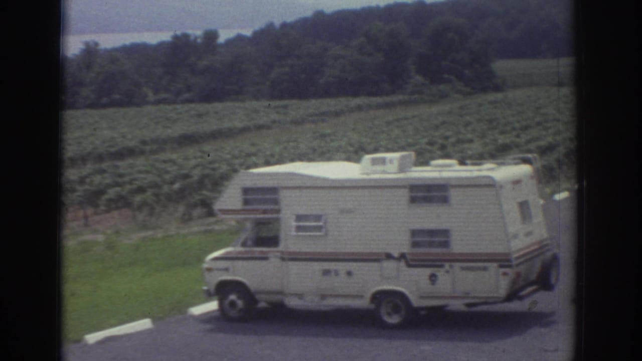 A white motorhome sits on a paved edge near green field rows, with wooded hills and a distant body of water visible in 1974 home-movie footage.