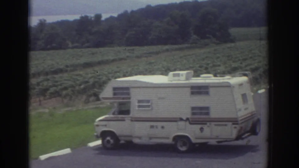 A white motorhome sits on a paved edge near green field rows, with wooded hills and a distant body of water visible in 1974 home-movie footage.