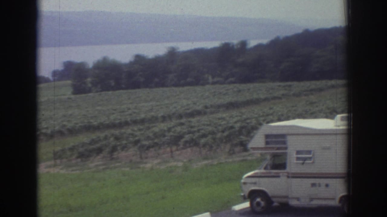 Partial view of the motorhome at right with field rows, trees, and distant water behind it.