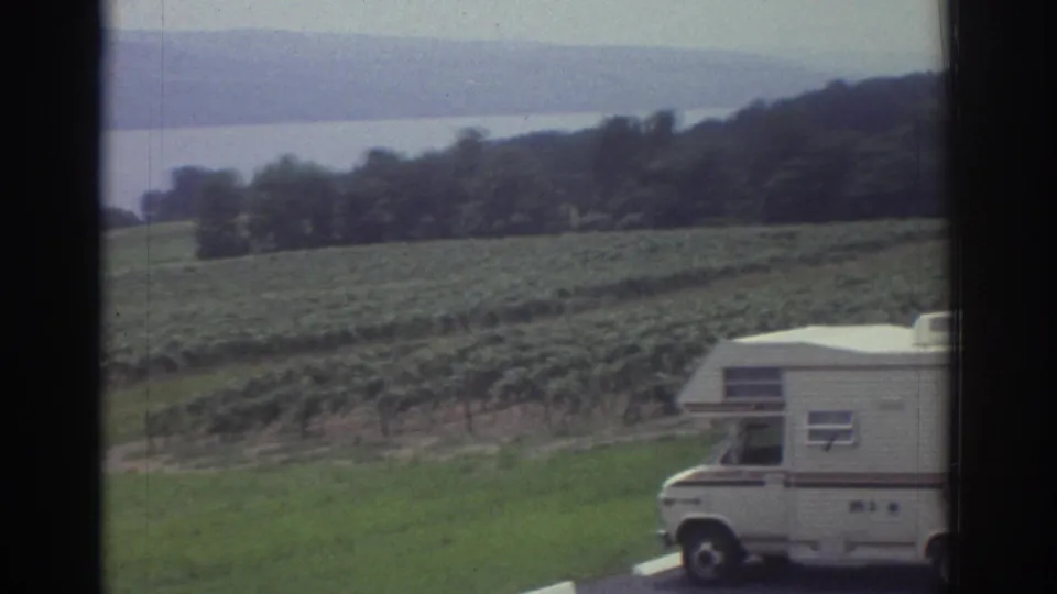 The motorhome is partly cropped at the right edge while the camera frames green rows, a wooded hill, and distant water near the travel stop.