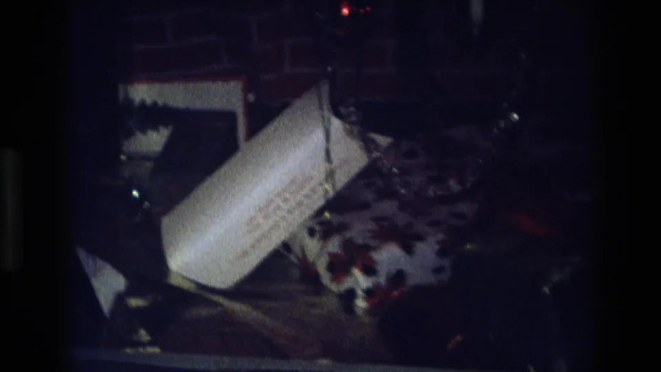 Dim 1975 Atlanta home-movie frame with a white plastic bag, patterned fabric, and household clutter near a brick wall.