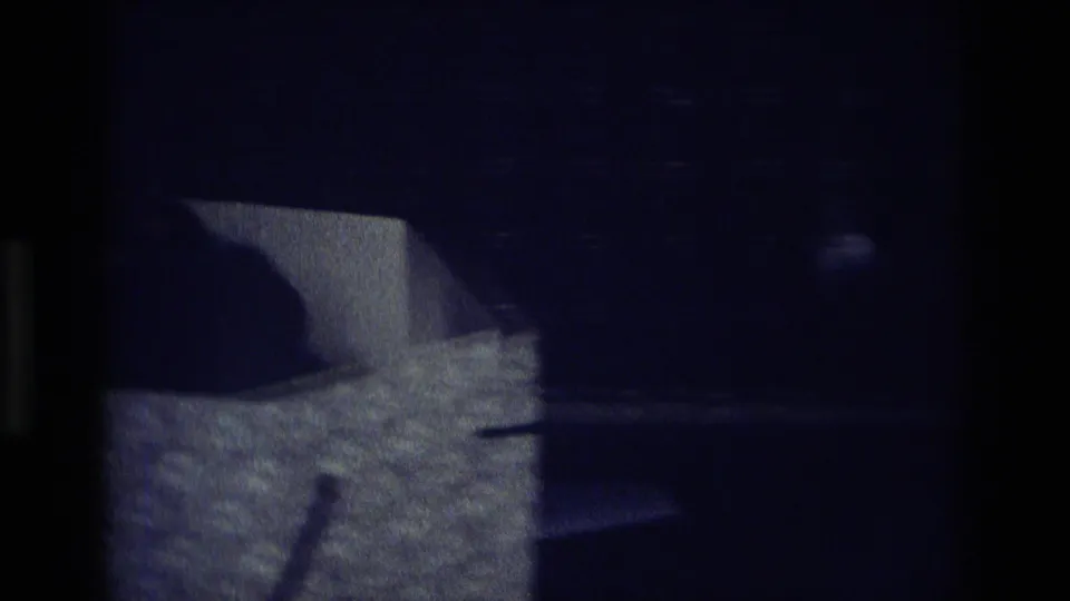 Underexposed 1975 Atlanta home-movie frame with a partial figure or object near a light-colored tabletop in a private interior.