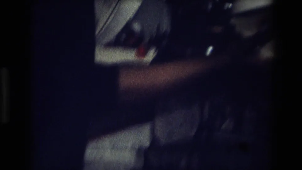 Low-light 1975 Atlanta home-movie frame showing a blurred partial person beside a white bag and dark shiny object in an interior setting.