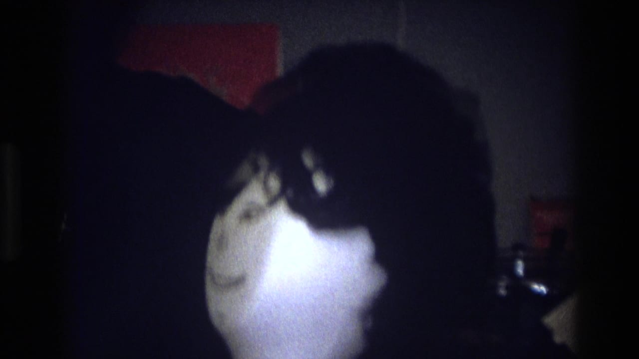 1975 Atlanta home-movie frame showing a bright-faced hair mannequin near a shadowed woman in a dark private interior.
