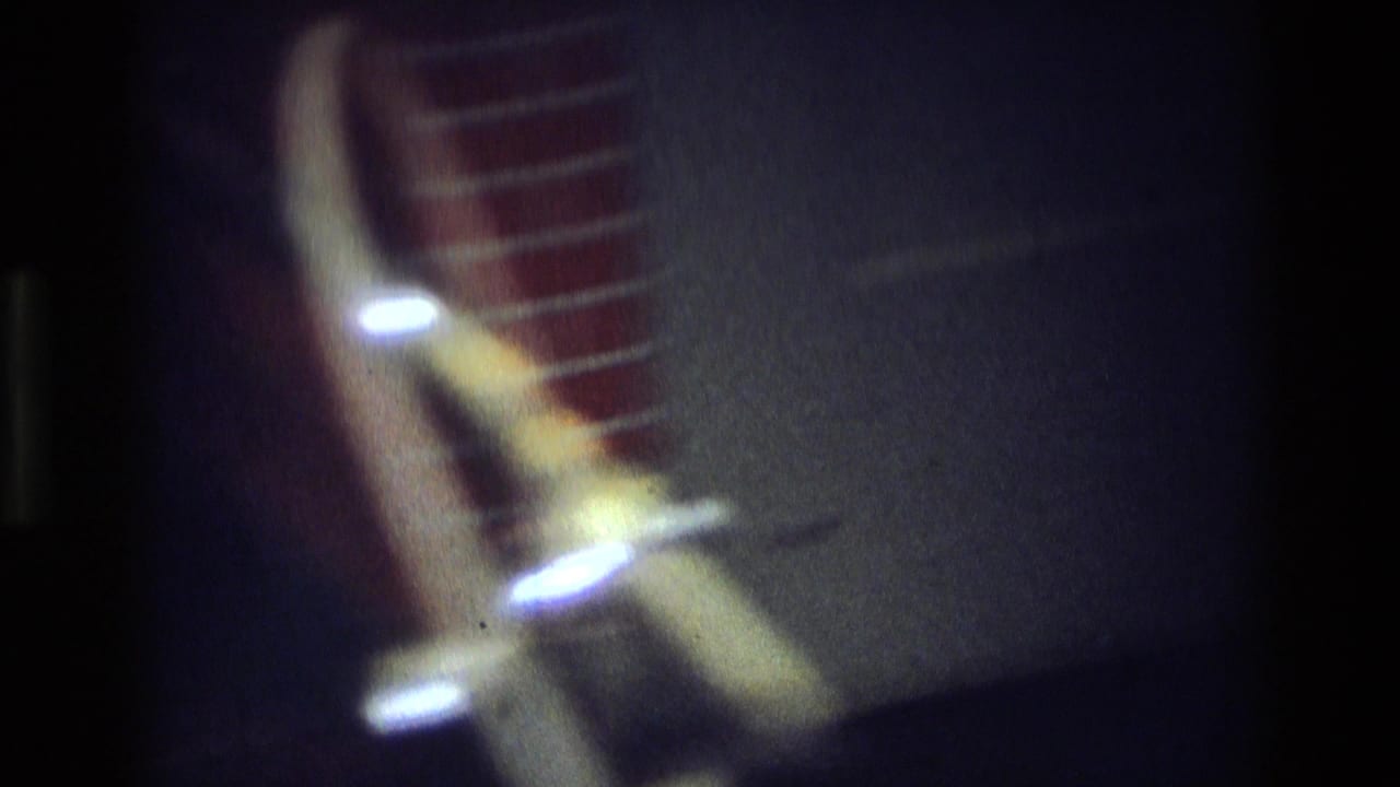 Soft, dark 8mm frame with blurred glowing lights and a red striped form, likely an interior or night transition shot.