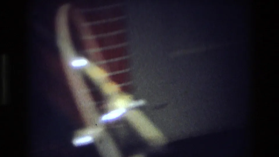 Soft, dark 8mm frame with blurred glowing lights and a red striped form, likely an interior or night transition shot.