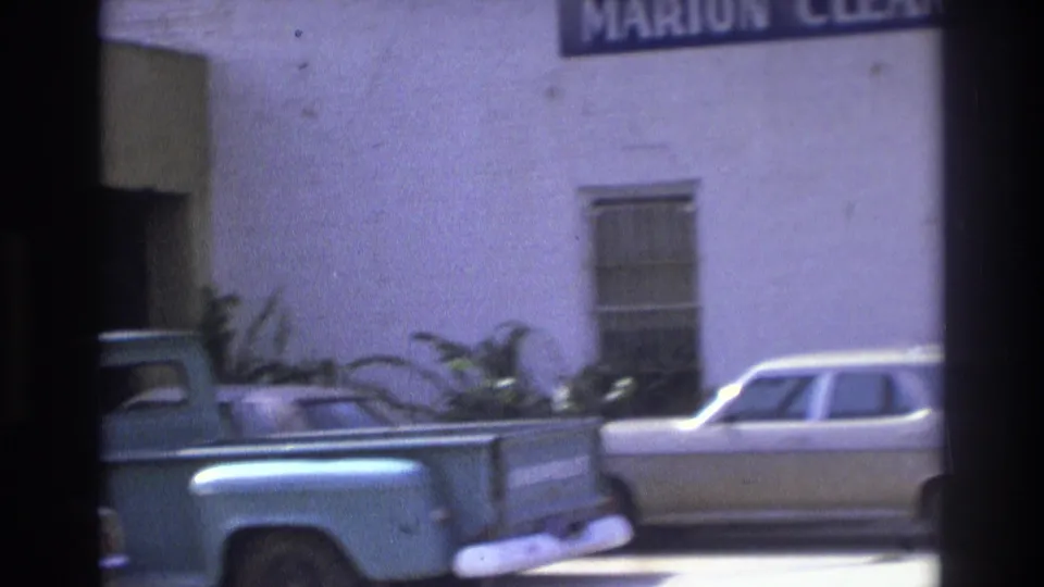 1975 Atlanta street frame with a light blue pickup, a pale sedan, plants, and a white commercial building with partial signage.