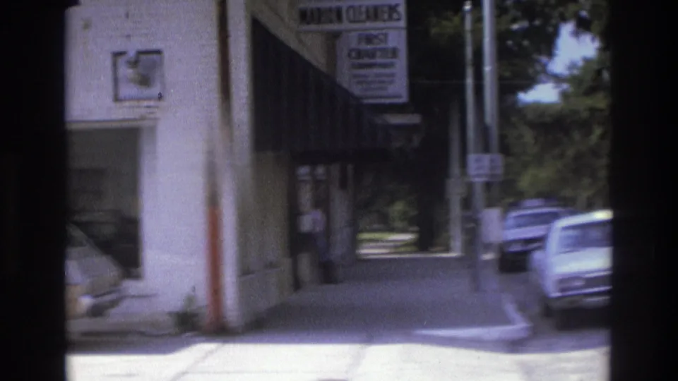 1975 Atlanta sidewalk view with storefront signage, a dark awning, parked cars, and a visible pedestrian near the doorway.