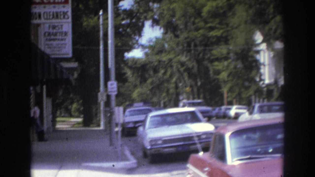 1975 Atlanta street scene seen from a moving vehicle, with parked cars, sidewalk, trees, and a partially visible business sign.