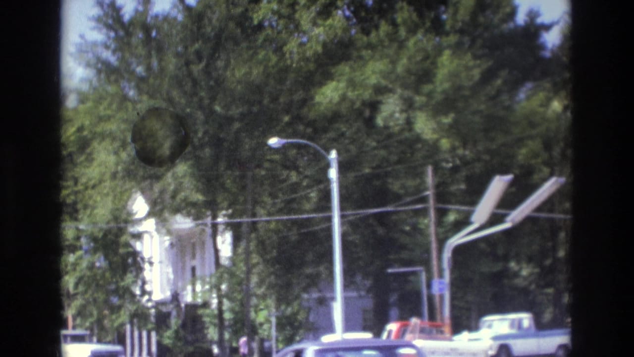 Tree-lined street with houses or small buildings, cars, streetlights, and dark film gate edges.
