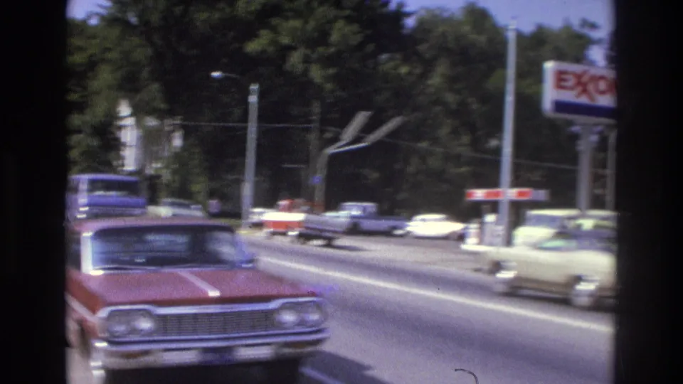 1975 Atlanta drive-by frame with a red sedan in front, moving traffic, and an Exxon sign near a roadside service station.