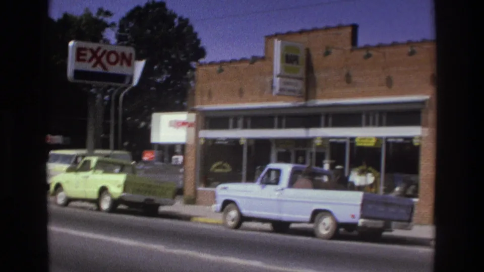 1975 Atlanta commercial street with brick storefronts, an Exxon sign, parked cars, and a light blue pickup truck at the curb.