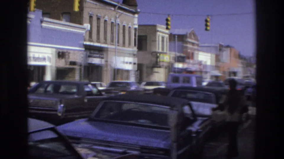 1975 Atlanta commercial block with multiple storefronts, curbside cars, overhead traffic lights, and a pedestrian passing near the camera.