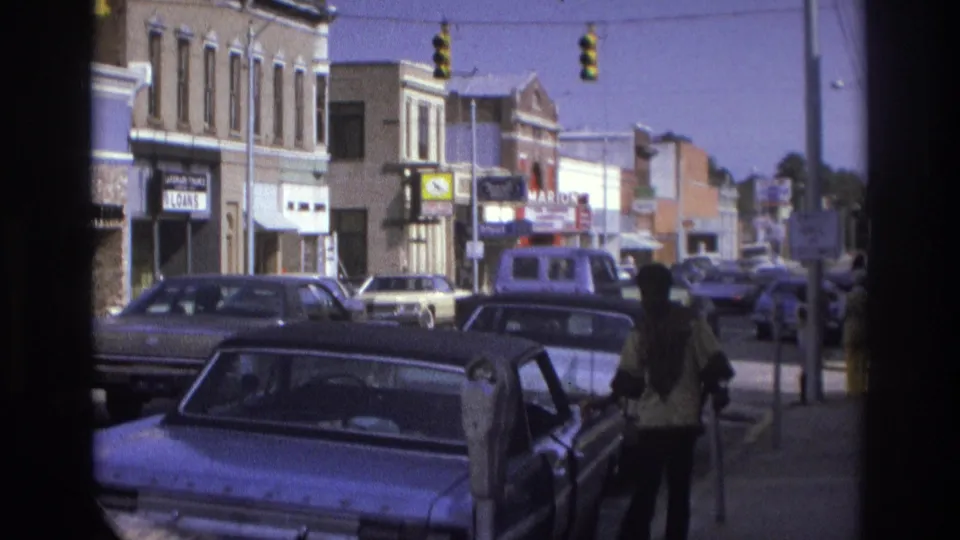 1975 Atlanta street view with classic cars, storefront signs, traffic signals, and a person standing near an open car door.
