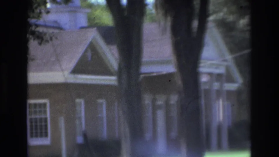 1975 Atlanta 8mm frame showing a gabled residential-style building partly hidden behind tree trunks and shade.