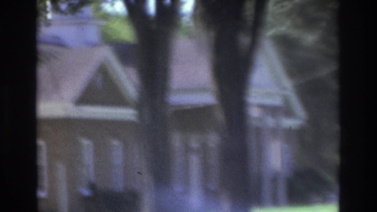 1975 Atlanta-area home-movie frame showing a pitched-roof house partly hidden by trees and dark vertical foreground obstruction.