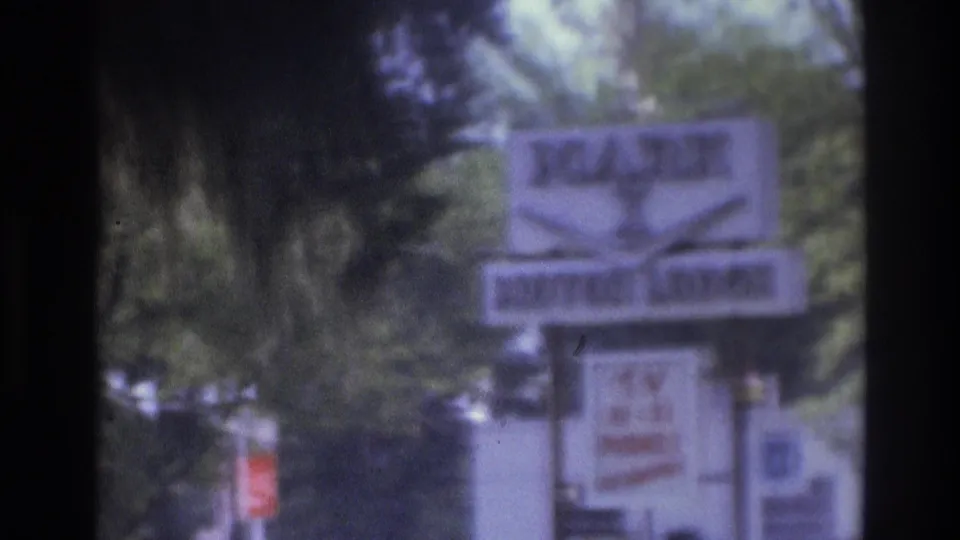 1975 Atlanta home-movie frame centered on a roadside camping or lodge-style sign, with trees and nearby structures in soft focus.