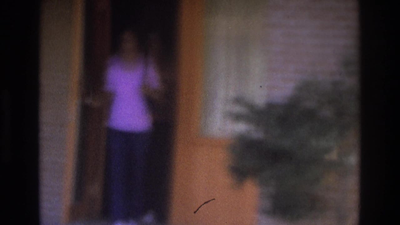 Atlanta 1975 home-movie frame with a blurred person in purple near a motel room doorway, seen through soft focus and dark frame edges.